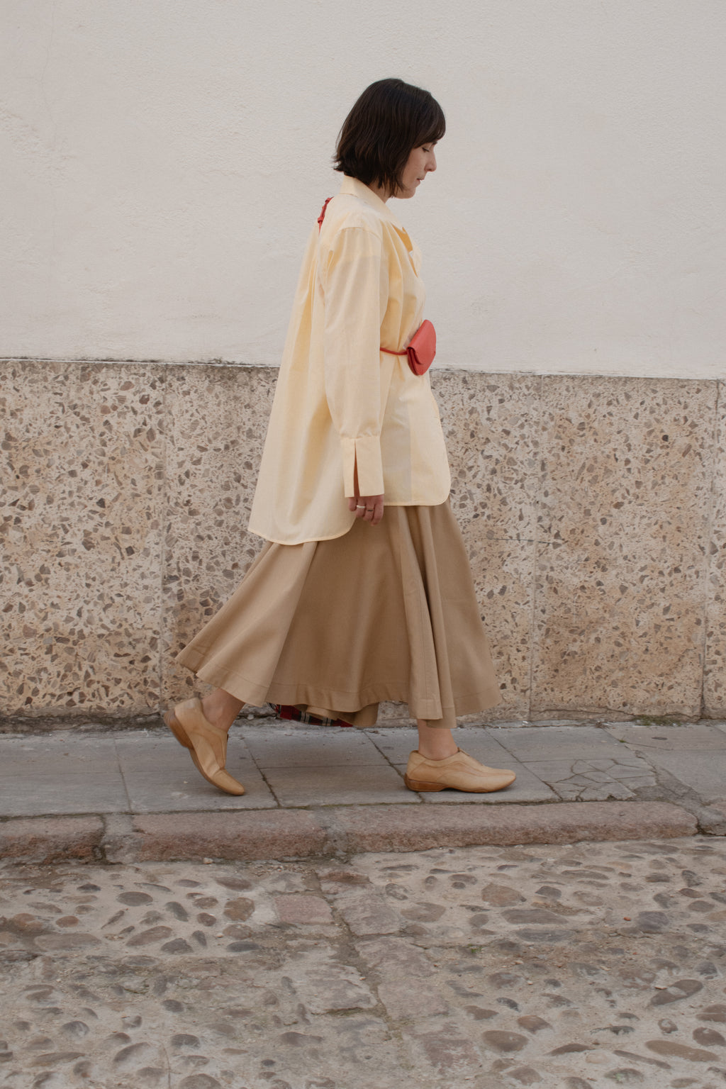 Woman walking on a stone pavement wearing a beige dress and light-colored shoes.