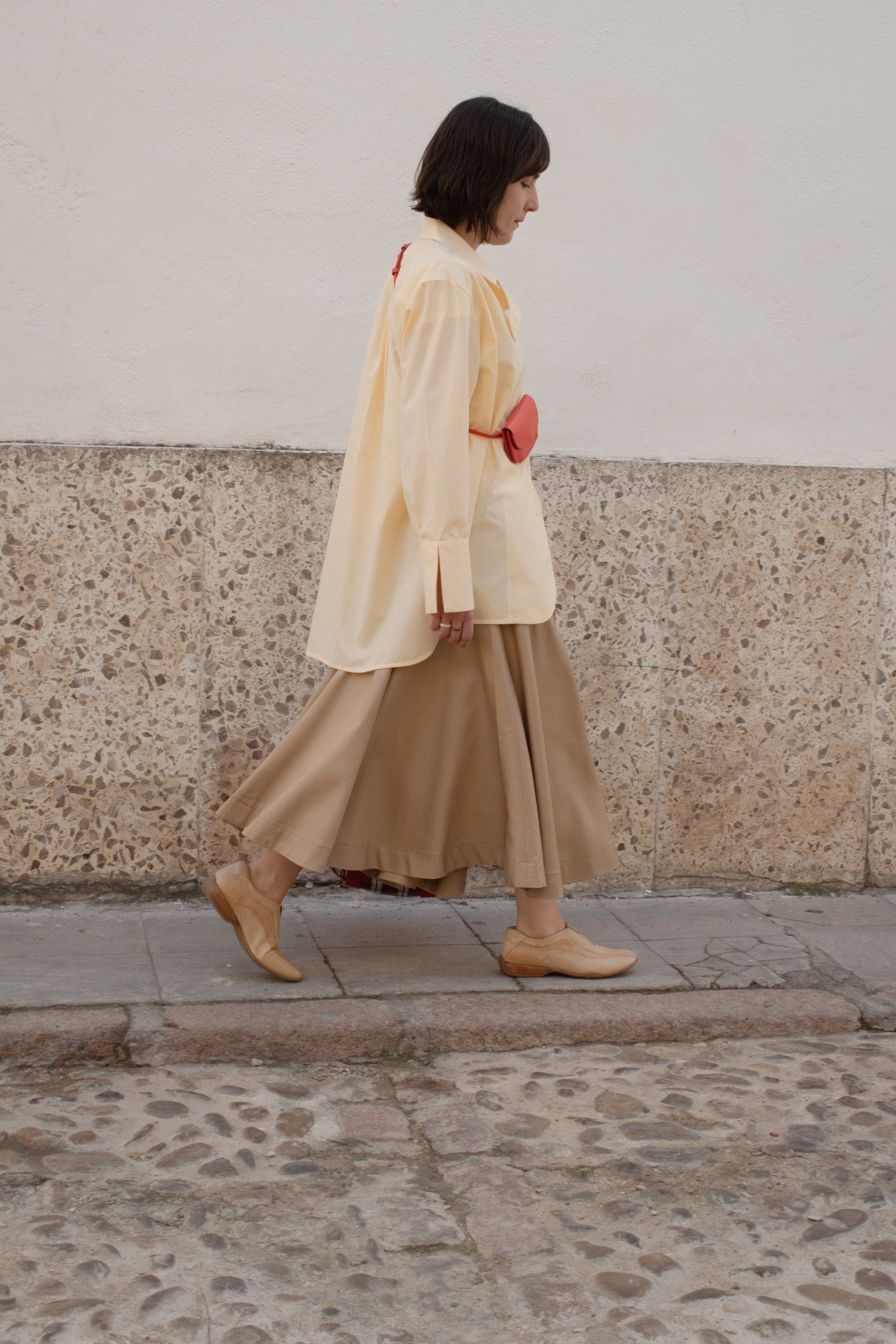 Woman walking on a stone pavement wearing a beige dress and light-colored shoes.